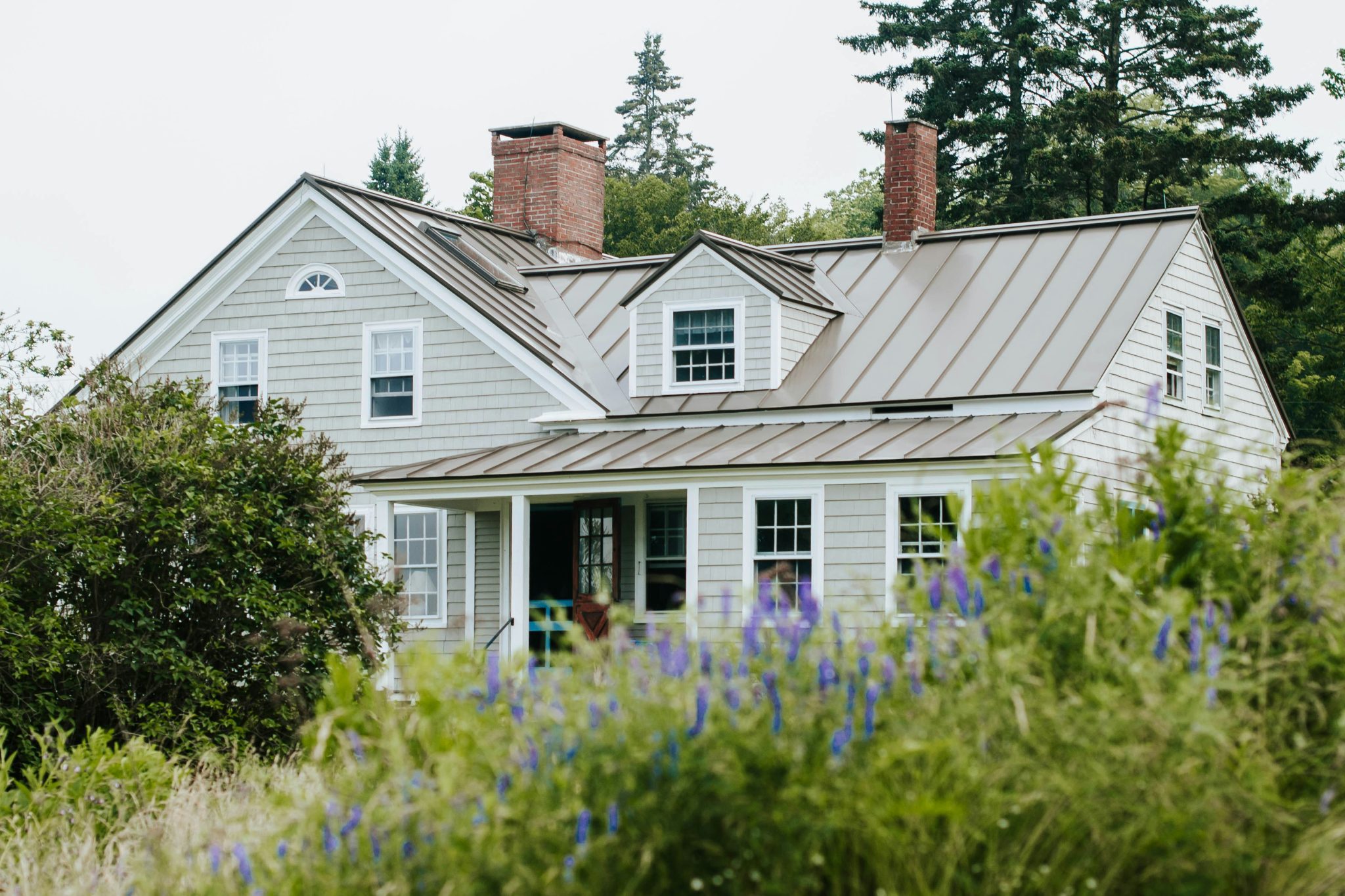Two-story house with standing-seam metal roof and chimneys, framed by shrubs and wildflowers — image for "Storm Damage vs. Wear and Tear."