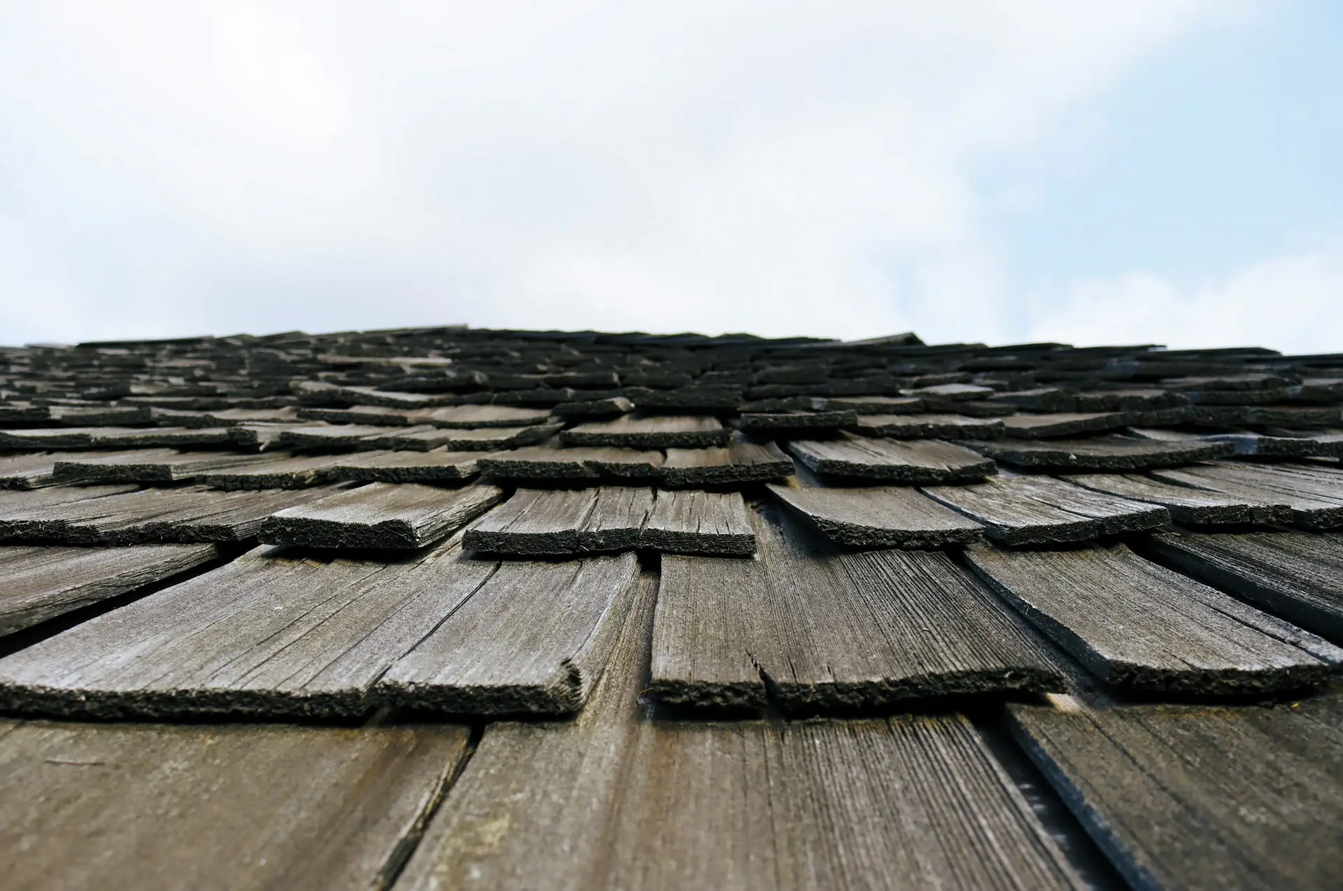 A low-angle, close-up view of weathered wooden roof shingles showing signs of wear and tear with graying wood and frayed edges, illustrating the subtle differences from sudden storm damage.