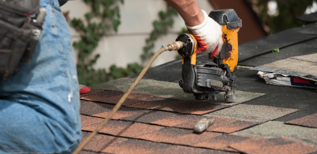 roofer using a pneumatic nail gun to install asphalt shingles