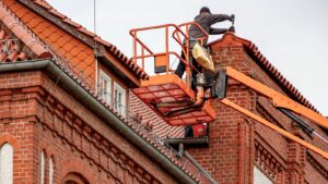Roofer in a cherry picker performing maintenance on a red brick building with a tiled roof.