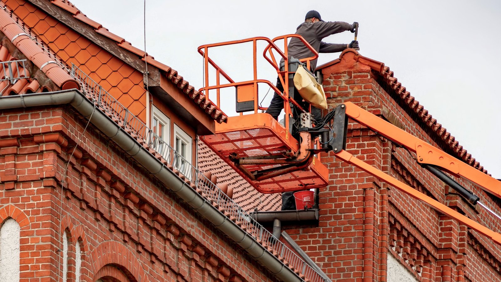 Roofer in a cherry picker performing maintenance on a red brick building with a tiled roof.